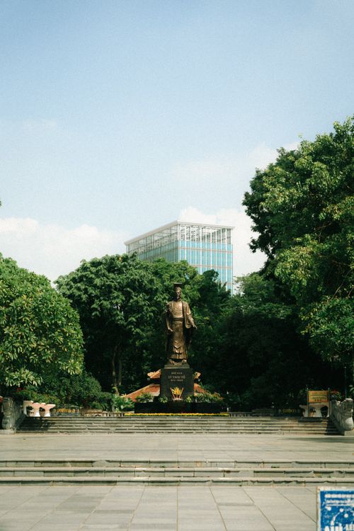 Free A serene statue stands in an urban park with lush greenery and a skyscraper backdrop under a clear sky. Stock Photo