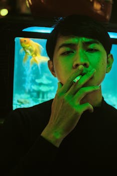 A contemplative man smoking indoors, illuminated by neon and aquarium glow, evoking a moody atmosphere.