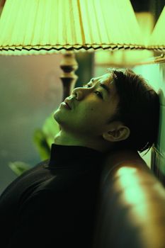 侧面图中，一位深思熟虑的亚洲男子坐在落地灯附近的皮沙发上