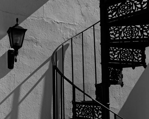 Free Artistic black and white photo of a vintage spiral staircase with intricate patterns and a wall lamp casting shadows. Stock Photo