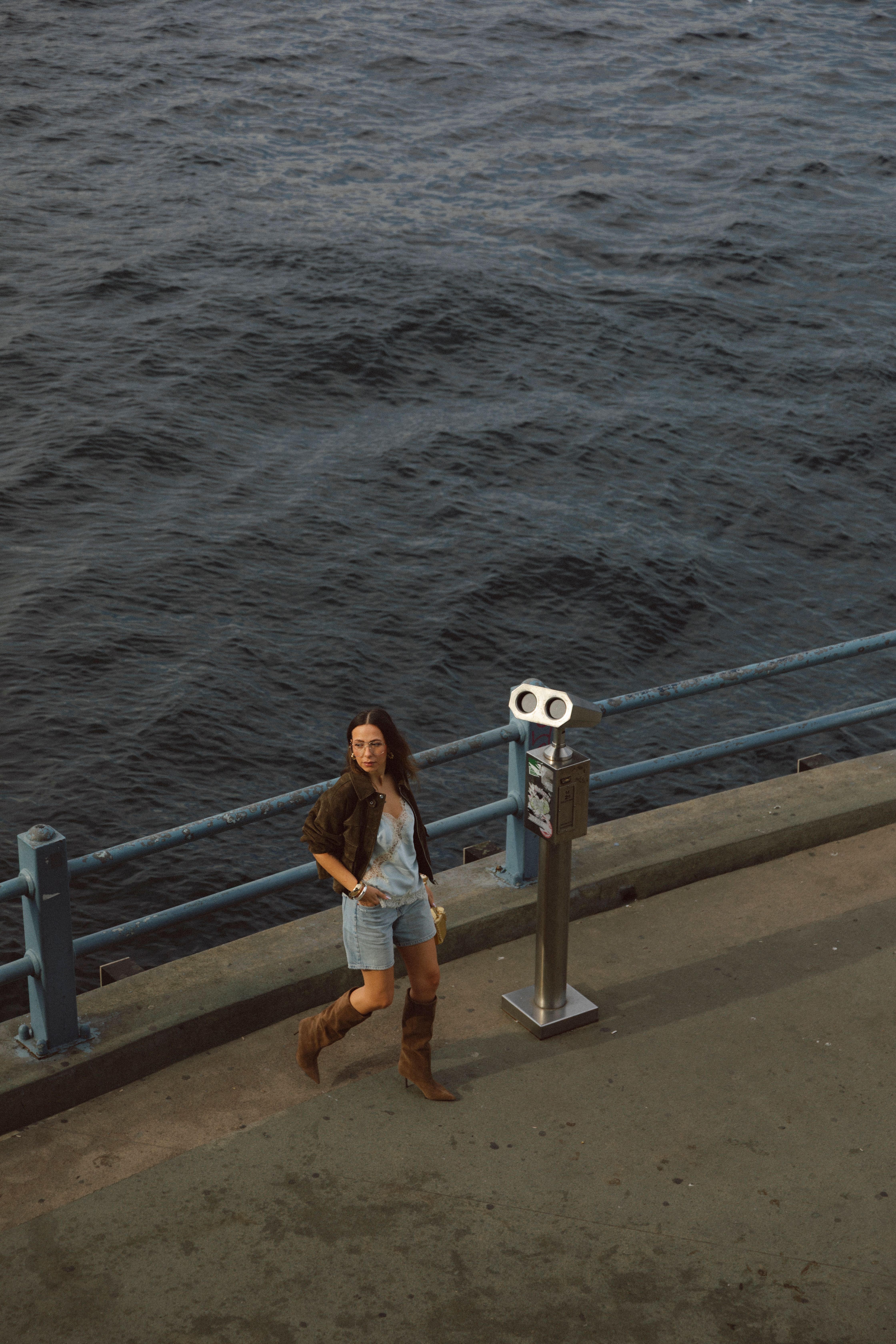 A woman stands near binoculars by the ocean railing, embracing the coastal breeze.