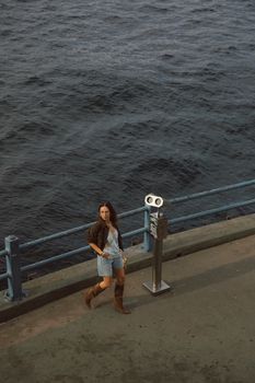 A woman stands near binoculars by the ocean railing, embracing the coastal breeze.