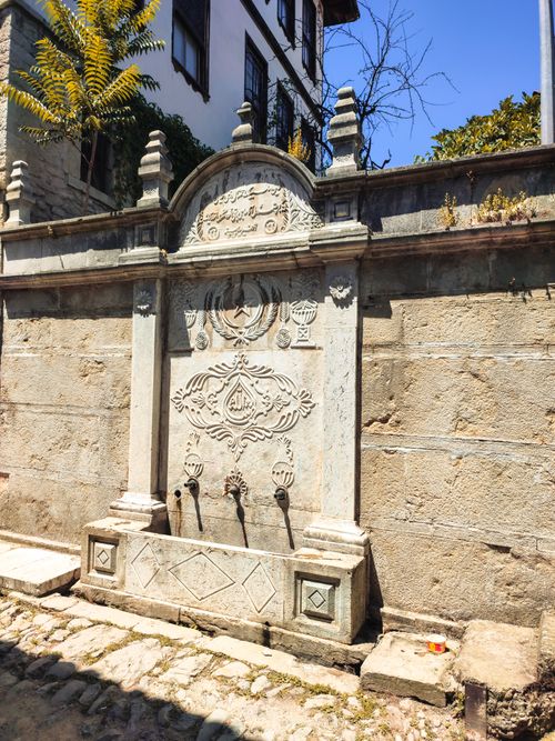 Free A detailed stone fountain with ornate carvings in a sunlit historic alley. Stock Photo