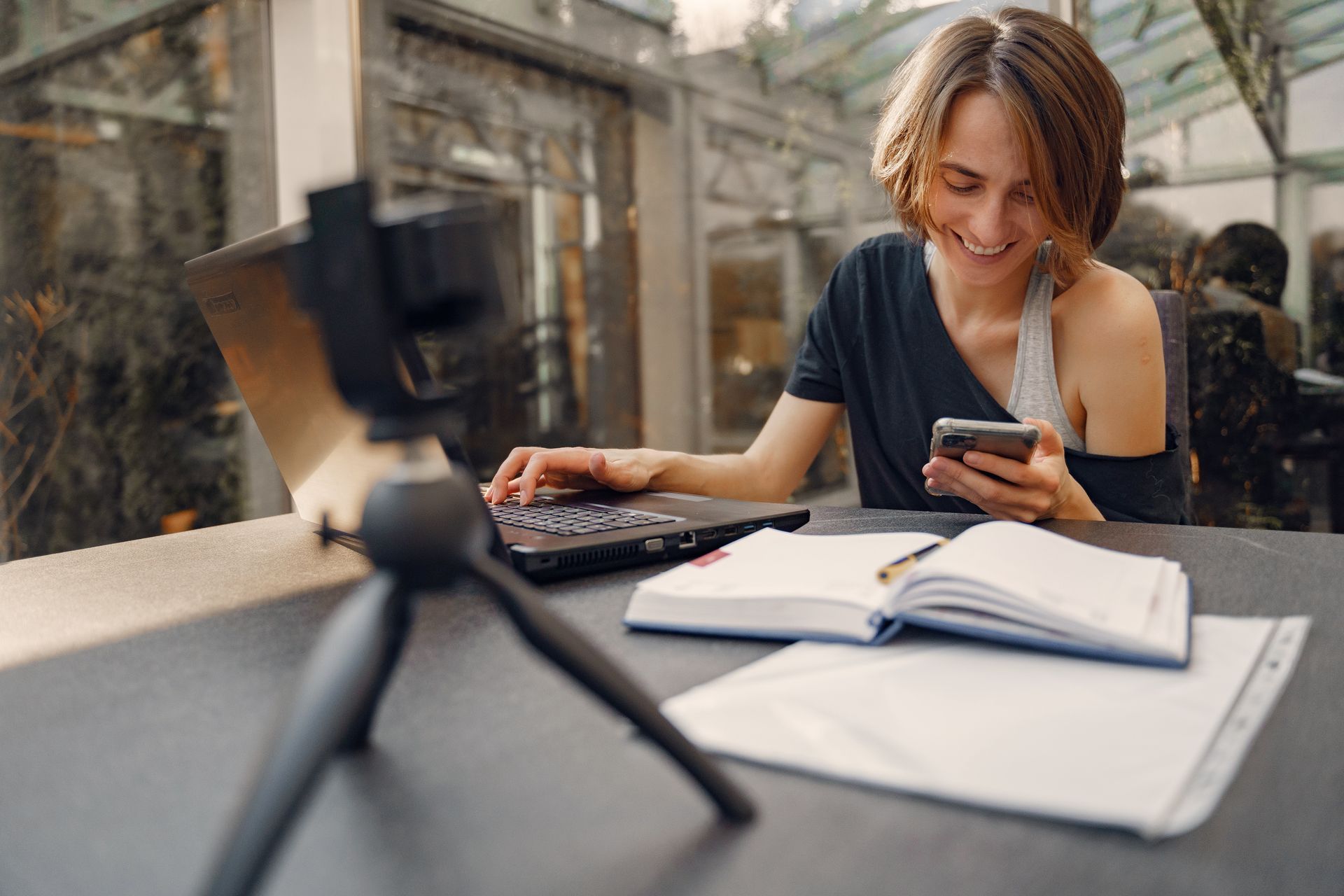 A woman smiles while working remotely with a laptop and smartphone at a desk.