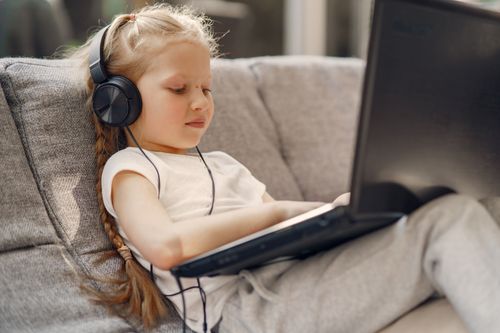 Free Small girl with long braid in headphones using laptop while sitting on sofa in living room Stock Photo