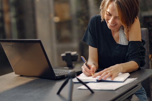 A woman smiling while working remotely, writing notes beside her laptop in a cozy indoor setting.