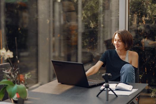 Woman enjoying remote work at home with laptop and notebook, smiling in a bright room.