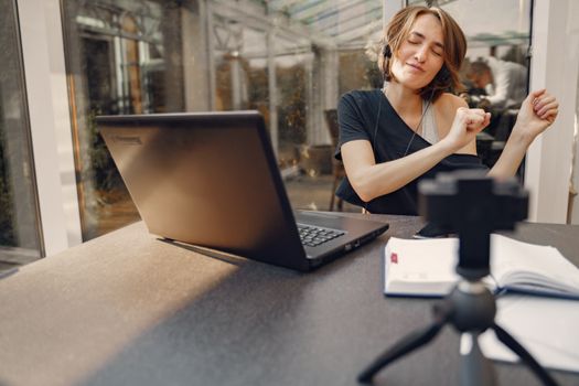 Cheerful female dancing while listening to music sitting with laptop and camera at table in office space