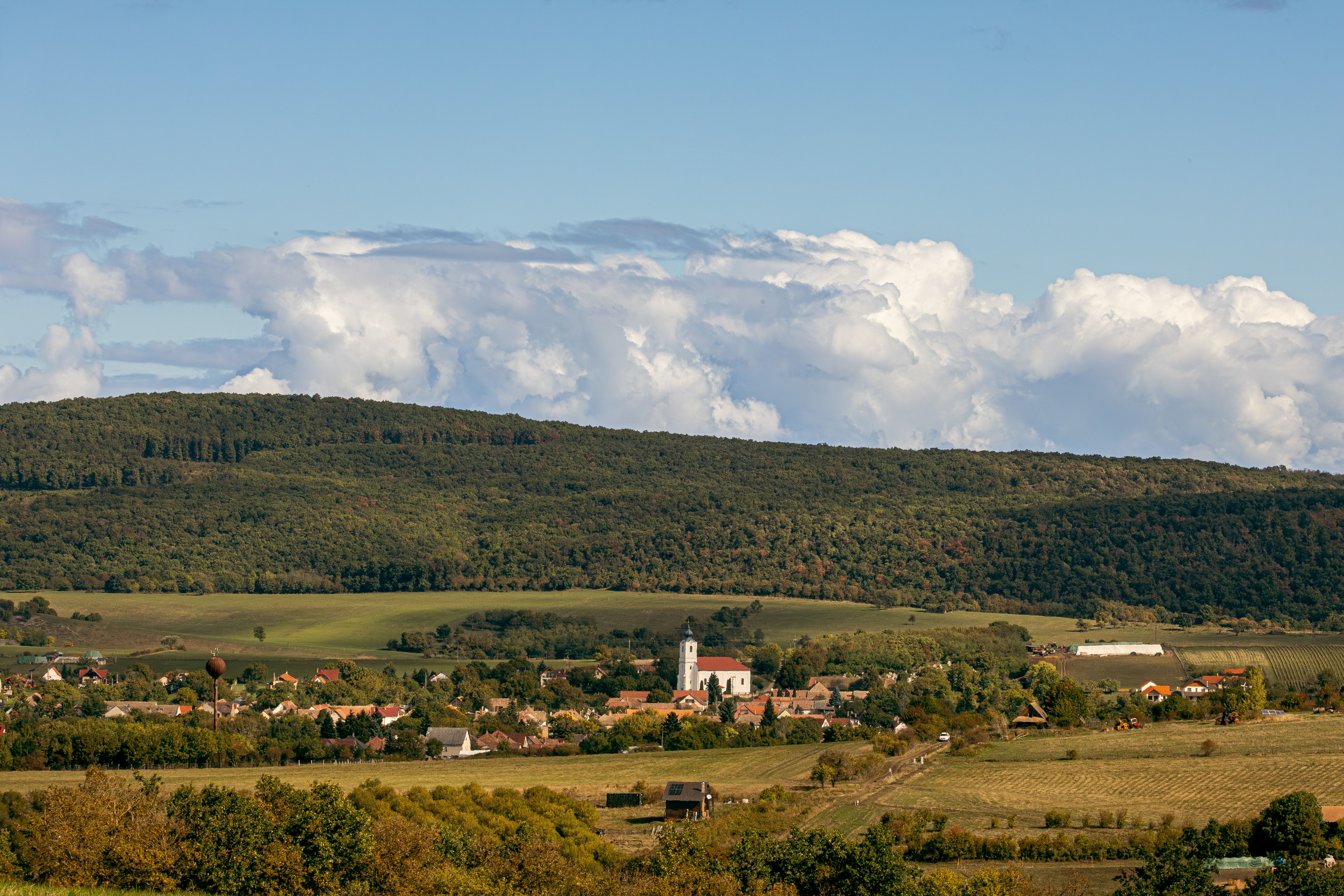 Scenic view of a Hungarian village surrounded by lush hills and cloudy sky, featuring a central church.