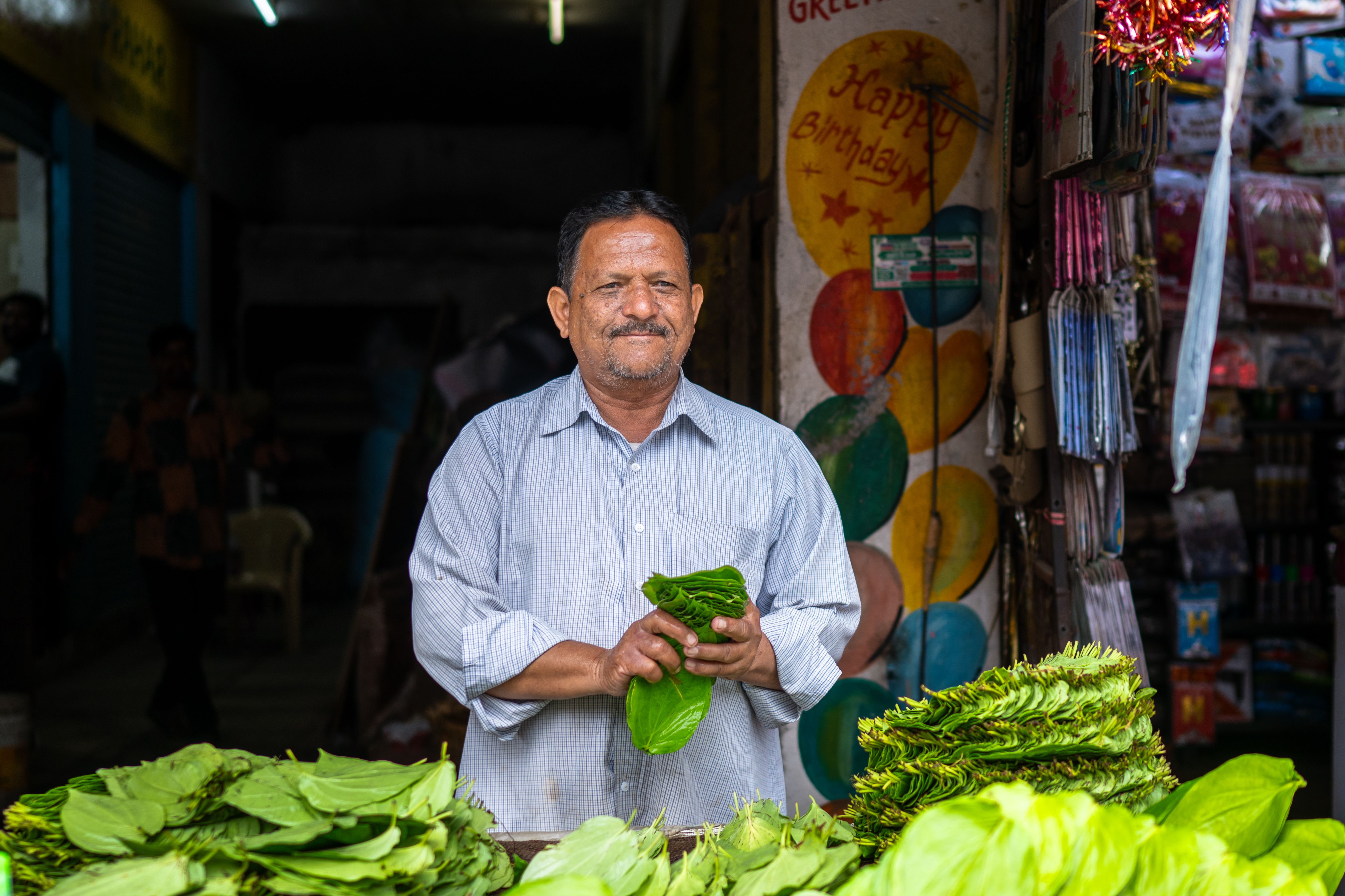 Smiling vendor holding fresh betel leaves at a vibrant market stall, perfect for travel and cultural themes.