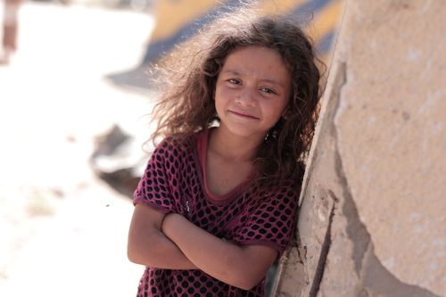 Free Smiling girl with curly hair leans against a wall in a sunny outdoor setting. Stock Photo