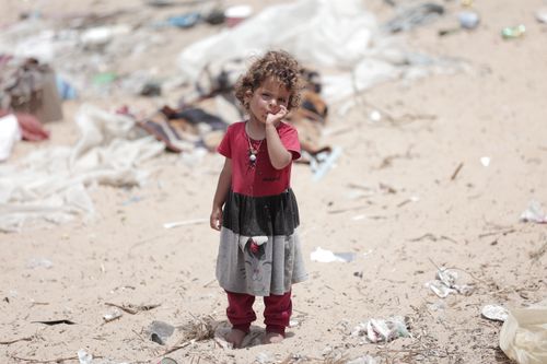 Free A young child stands on a sandy beach surrounded by discarded debris, looking thoughtful. Stock Photo