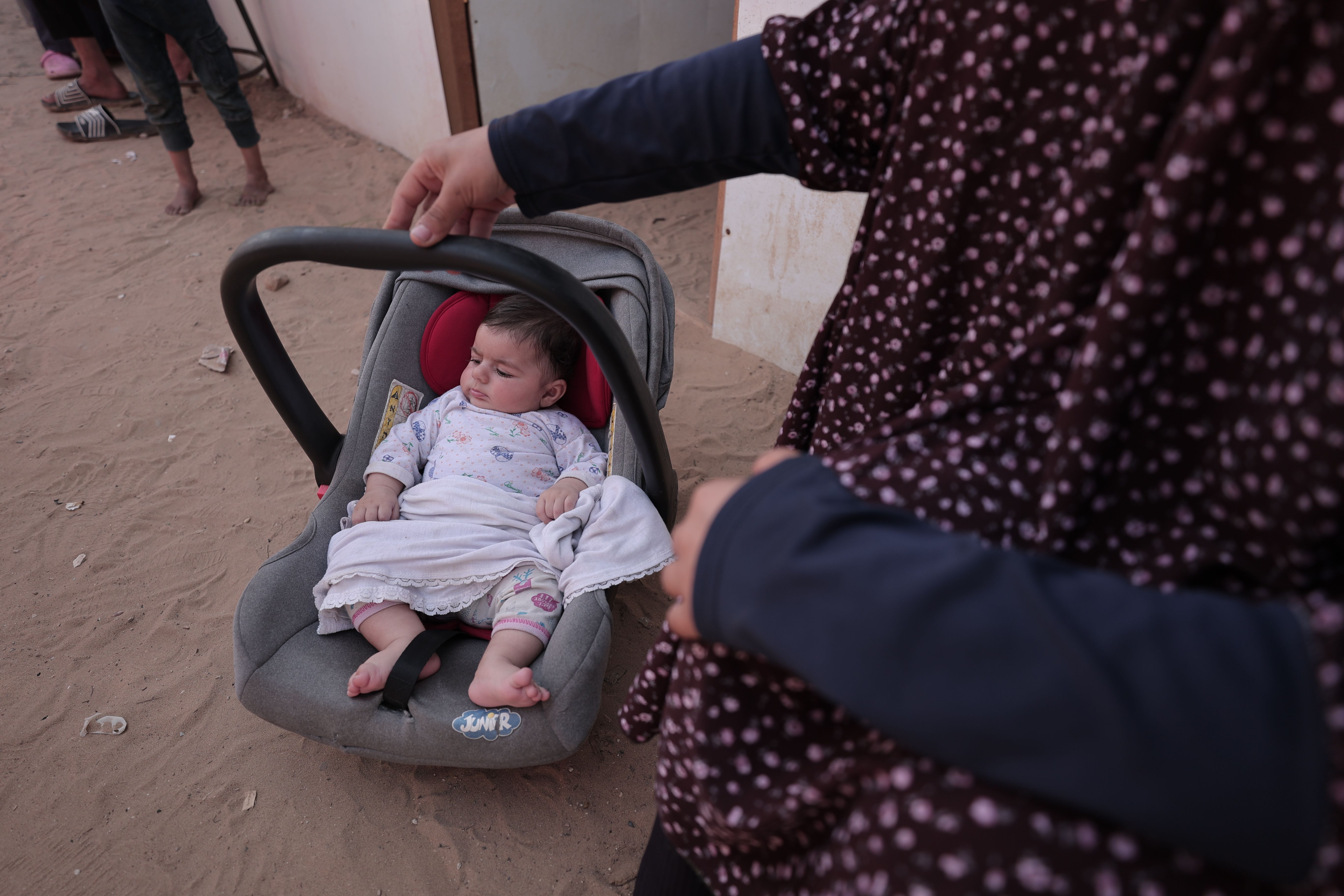 A baby sleeping in a car seat outdoors, held by a woman dressed in floral attire, creating a peaceful scene.