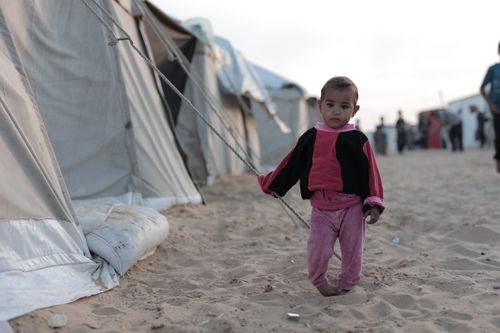 Free A young child in pink clothing stands in a sandy refugee camp next to tents. Stock Photo