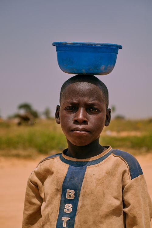 Free Portrait of a young African boy balancing a blue bowl on his head in a rural area of Katsina, Nigeria. Stock Photo