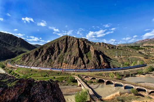 A train travels through the picturesque mountains of Sivas, Turkey, around a historic bridge.