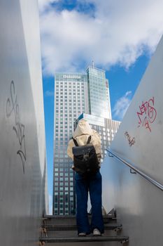 A person ascends a stairway with a view of a modern skyscraper under a blue sky in Rotterdam.