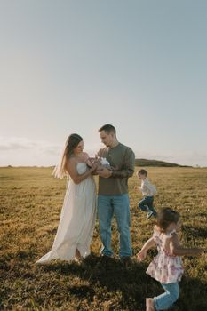 A family enjoys a sunny day in the field, celebrating with their newborn child and playful kids.