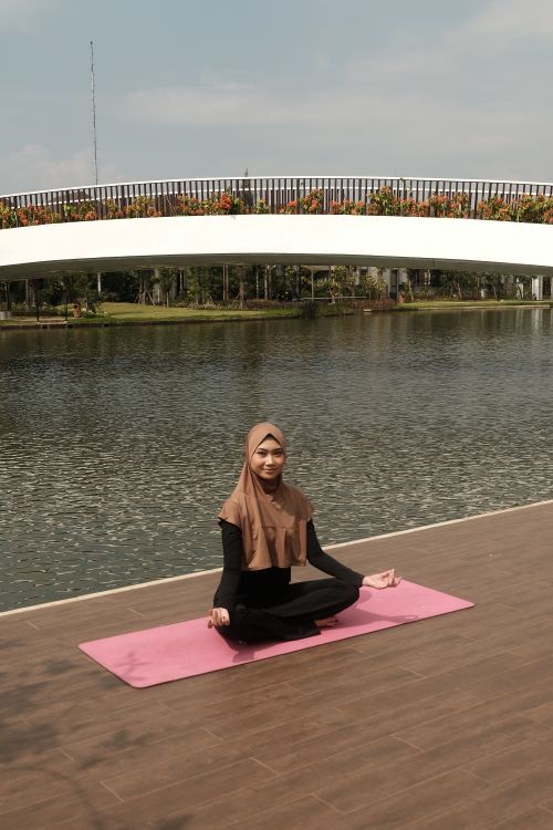 Free A woman practicing yoga on a pink mat near a lake with a curved modern bridge in the background. Stock Photo