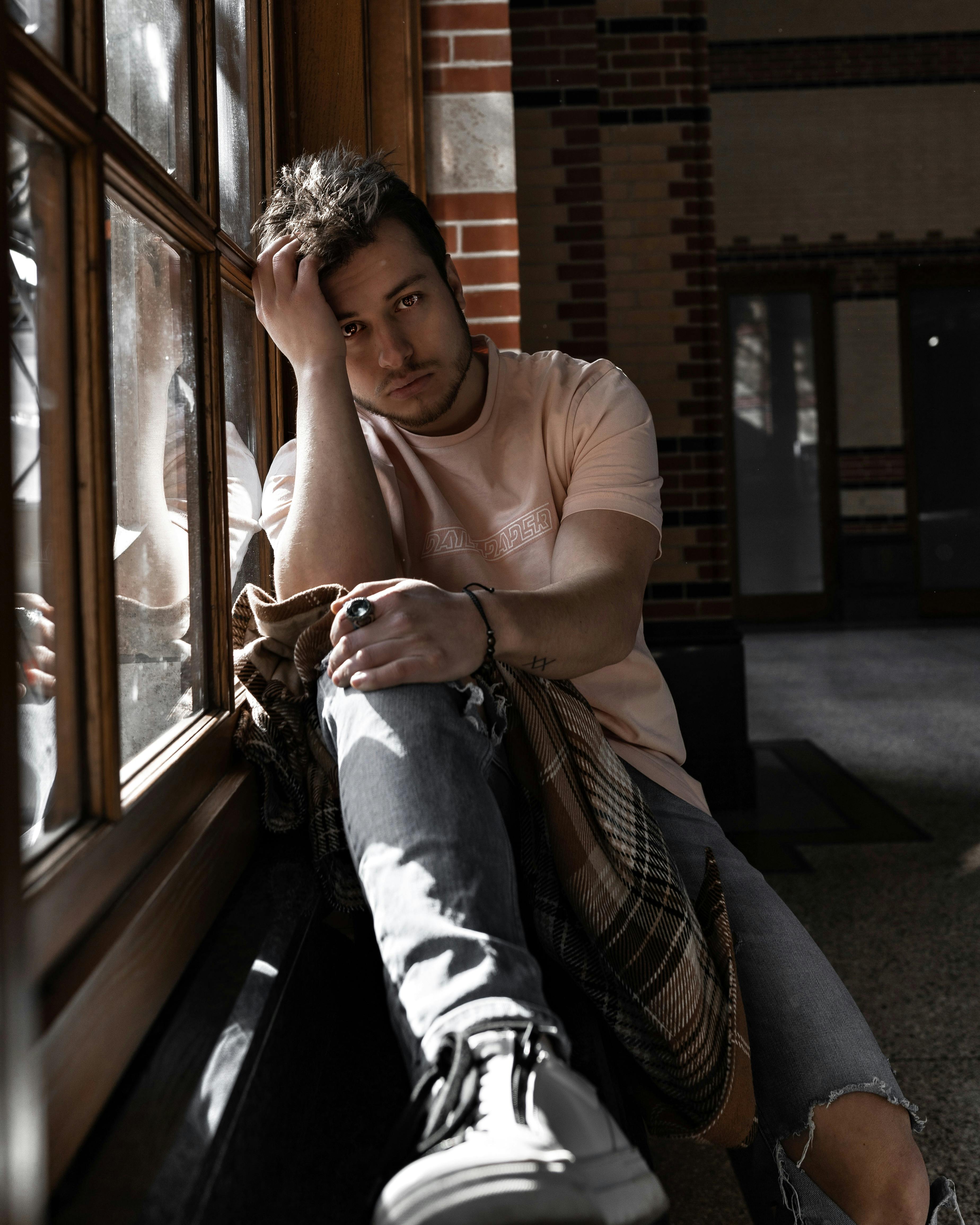 一个穿着休闲装、沉思的年轻人坐在室内窗前，反思着自己的想法