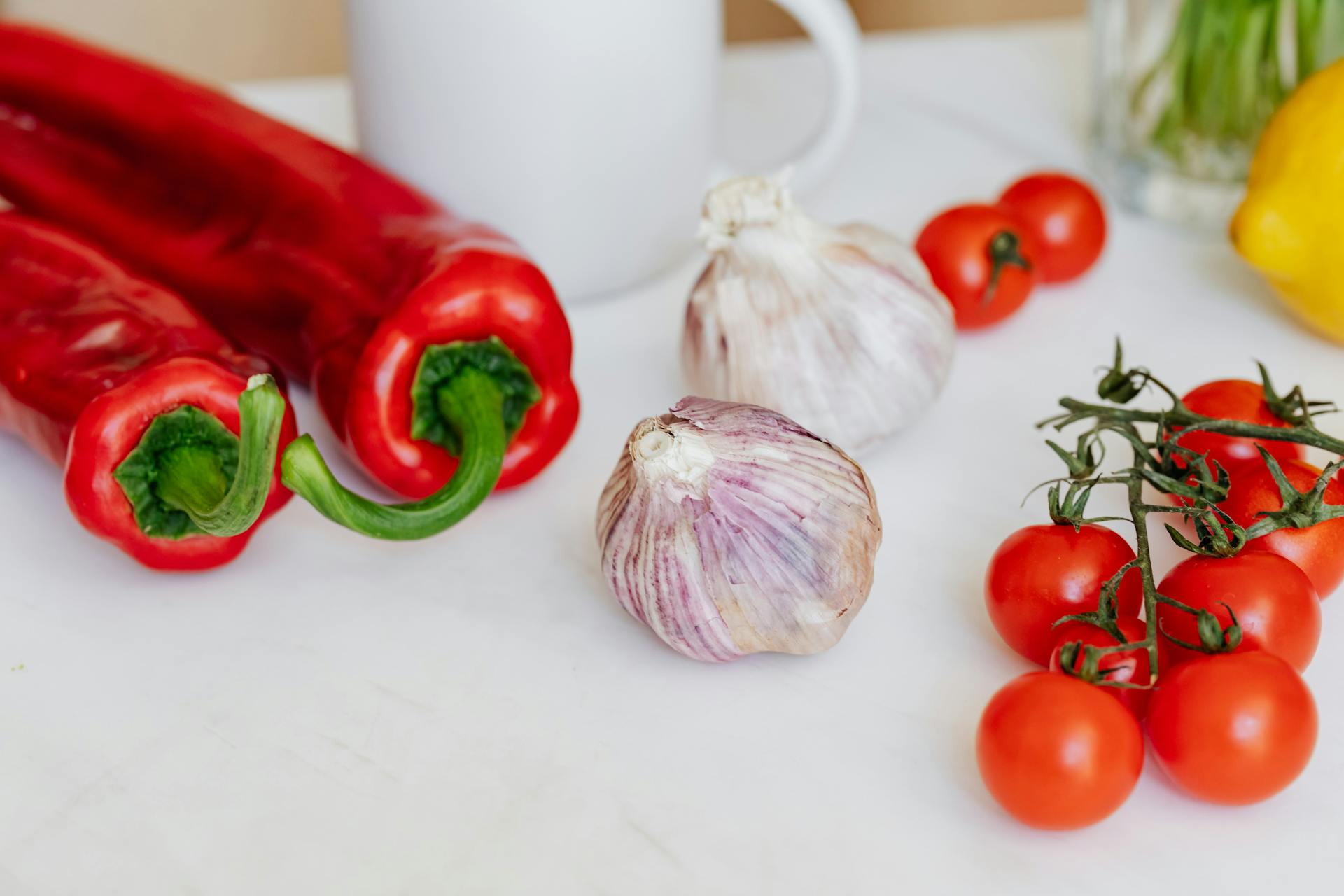 Fresh garlic placed among ripe red cherry tomatoes and peppers on white table near white ceramic cup and yellow lemon