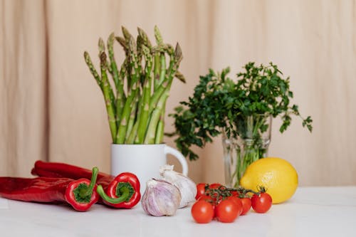 Composition of red cherry tomatoes and peppers with garlics and yellow lemon near white cup with asparagus and green parsley in glass on table