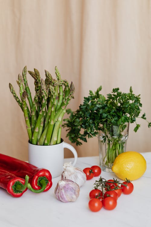 Bright red chili peppers and cherry tomatoes placed on table near yellow lemon and garlics against cup with asparagus and green bunch of parsley in glass on table