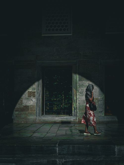 Free A woman walks through a dimly lit urban passage, highlighted by dramatic shadows. Stock Photo
