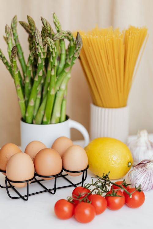 Metal stand with eggs among colorful vegetables and lemon placed on white table against green asparagus and spaghetti in white cups