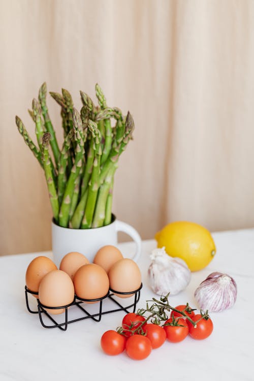 Free Bunch of red cherry tomatoes arranged before green asparagus and yellow lemon near garlics and metal basket with eggs on table Stock Photo