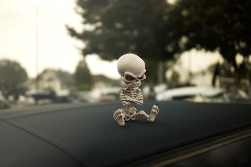 Free A whimsical skeleton figurine sits on a car roof with a blurry city background, enhancing its playful vibe. Stock Photo