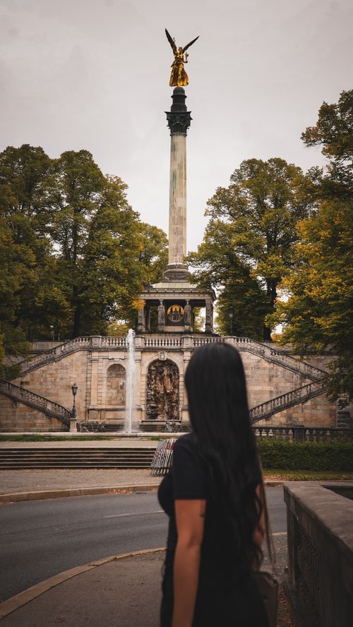 Free A woman stands before the Angel of Peace monument in Munich, surrounded by lush trees. Stock Photo