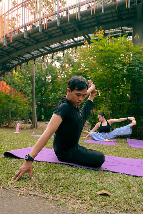 Free People practicing yoga outdoors in a tranquil park setting under a bridge, highlighting relaxation and wellness. Stock Photo