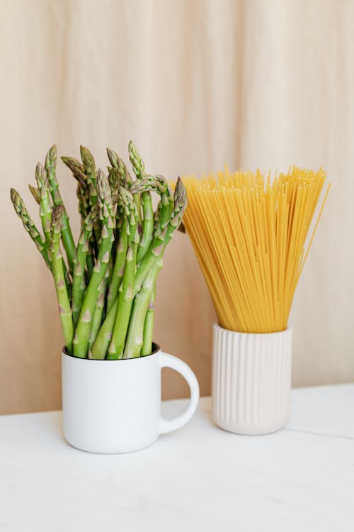 Piles of fresh green asparagus placed on white table near uncooked spaghetti in vase in light studio