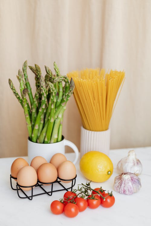 Various fresh vegetables and yellow lemon placed on white table near spaghetti in ceramic glass and raw eggs prepared for cooking pasta