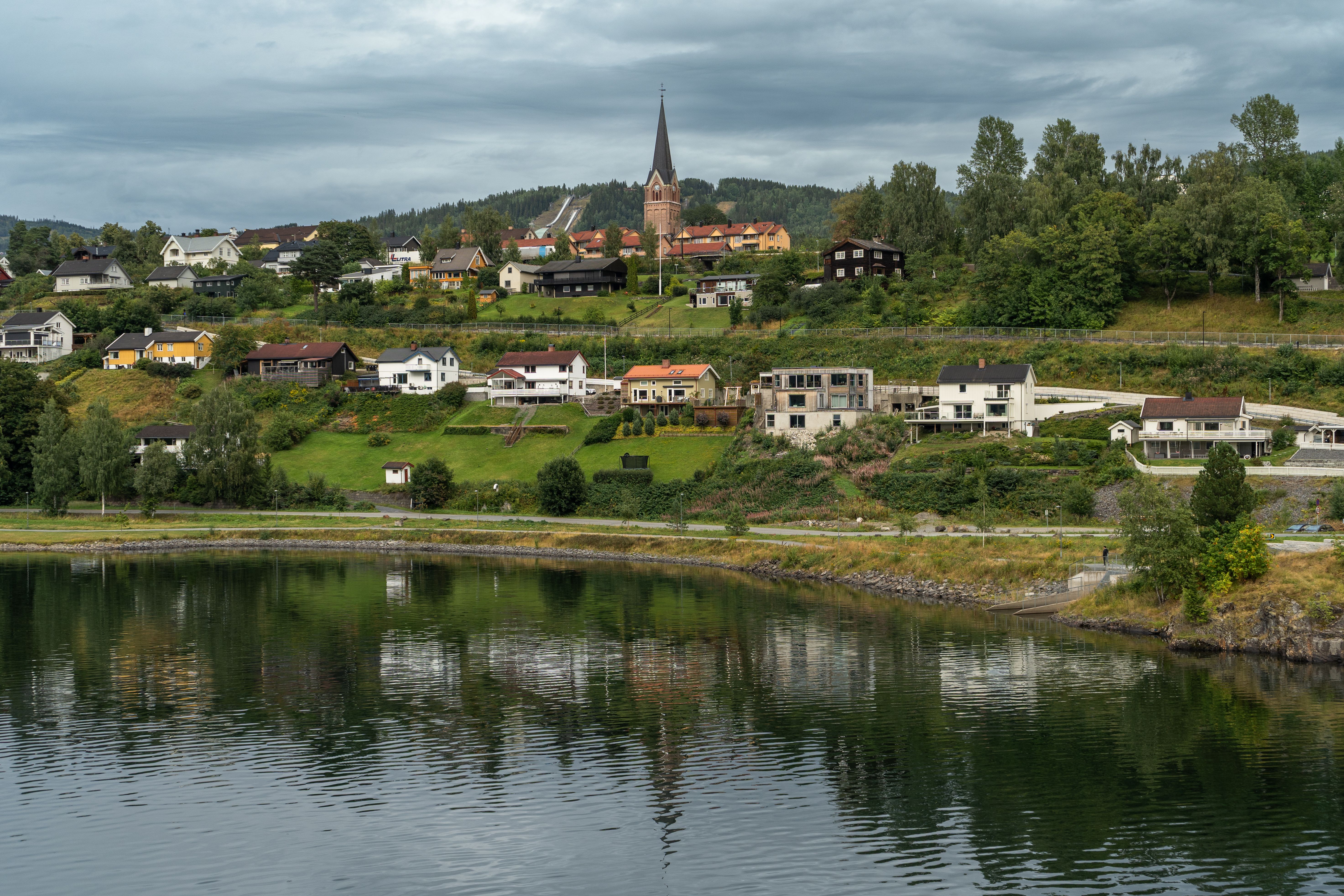 A picturesque Norwegian village with a church, reflecting on a calm river under a cloudy sky.