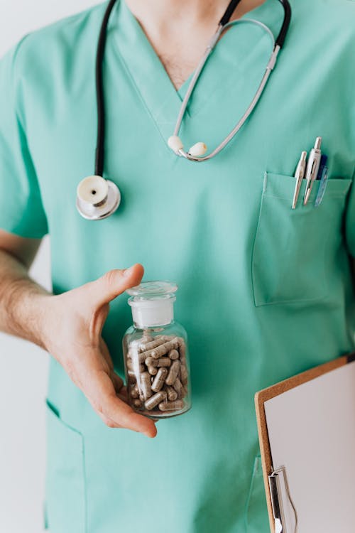 Faceless crop male doctor in medical uniform holding clipboard and demonstrating bottle with drugs while standing with stethoscope and pens in pocket