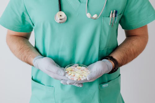 Unrecognizable male doctor wearing medical uniform and latex gloves holding Petri dish filled with assorted drugs while standing near white wall in hospital