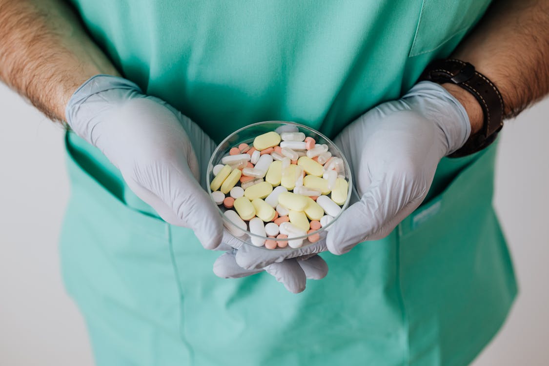 Crop male doctor wearing medical uniform and latex gloves holding glass bowl with colorful different drugs with crossed hands during work