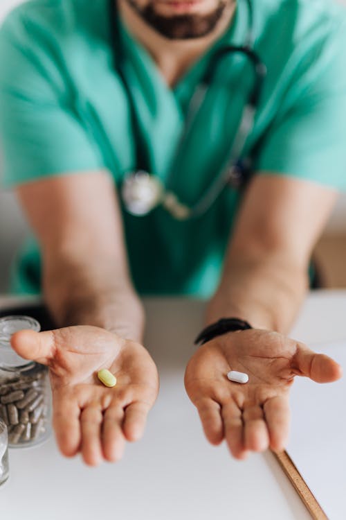 Crop man doctor wearing medical uniform demonstrating medicine to patient in clinic during work sitting at table with stethoscope and clipboard