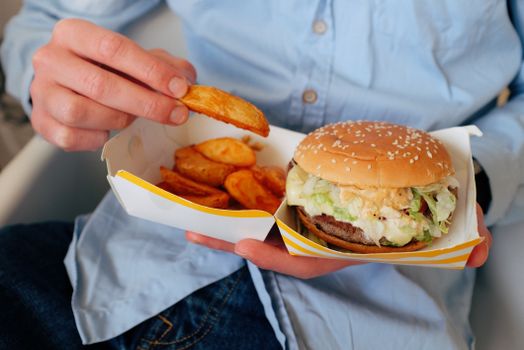 Anonymous male  in casual clothes eating tasty fast food consisting of burger and french fries while sitting on chair