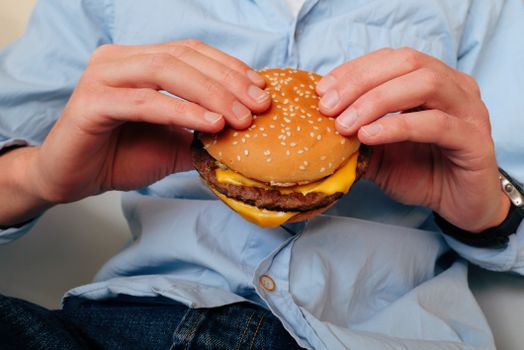 Crop male in casual wear holding burger consisting of cheese and cutlet while sitting on chair