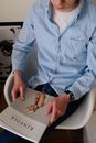 Crop smiling male with wristwatch in blue shirt and jeans sitting on chair and holding art magazine in hands in room