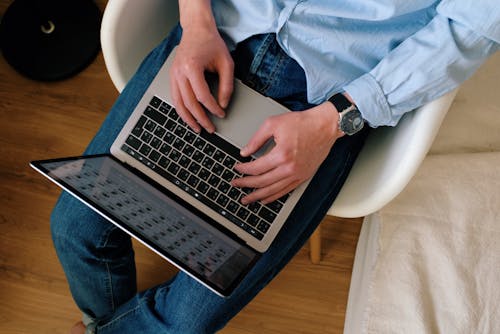 Top view of crop anonymous man in casual outfit using laptop and typing on keyboard while sitting on chair in modern apartment