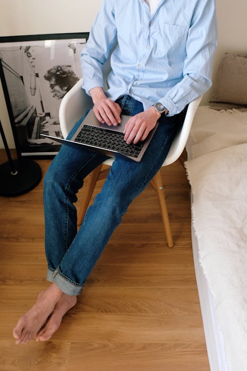 From above of young man in casual clothes sitting on chair in modern bedroom while typing on keyboard of laptop