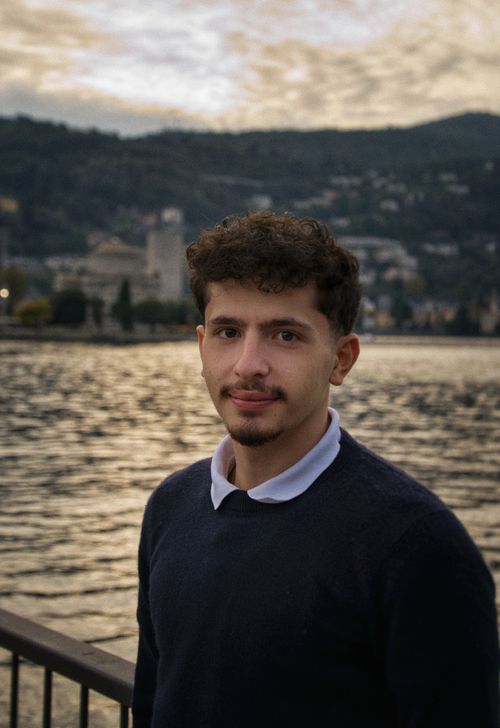 Free A young man stands by a tranquil lakeside with hills in the background during sunset. Stock Photo