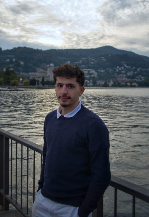 Free A young man stands by a waterfront with scenic mountains in the background during twilight. Stock Photo