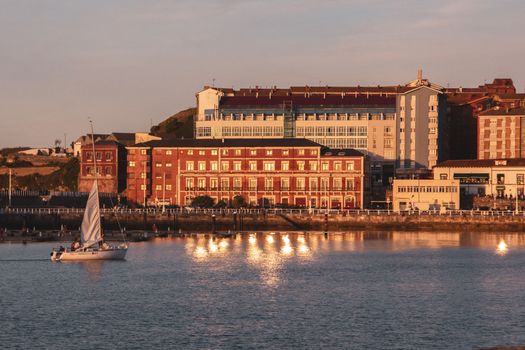 A serene sailboat in Gijón harbor during a picturesque sunset, capturing the city's historic architecture and tranquil waters.