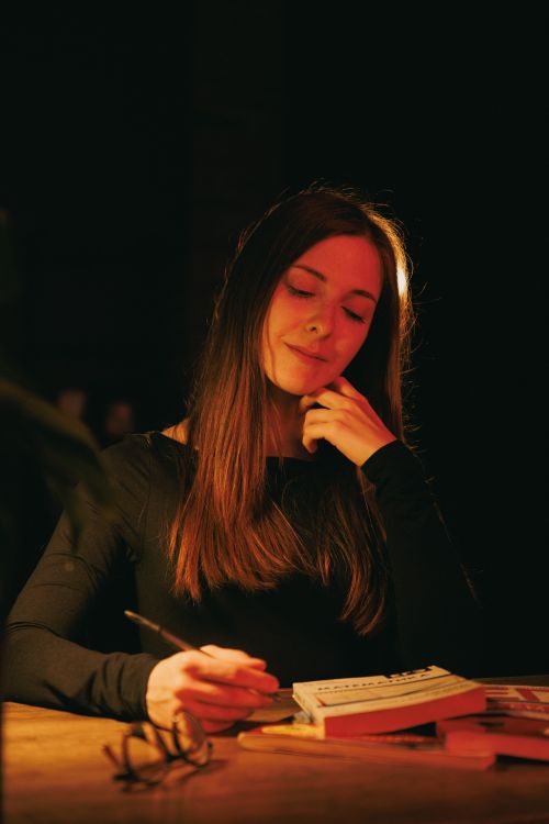 Free A woman in warm light pens thoughts at a desk, surrounded by books. Stock Photo