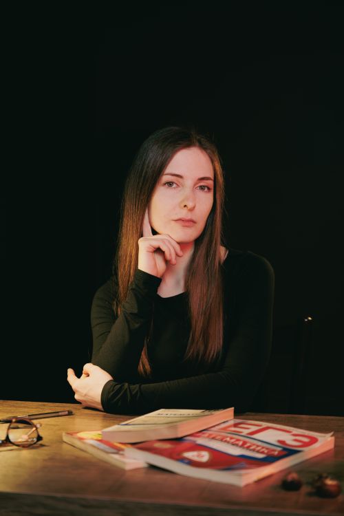 Free A thoughtful woman sitting at a table with books and glasses, contemplating in a dark setting. Stock Photo